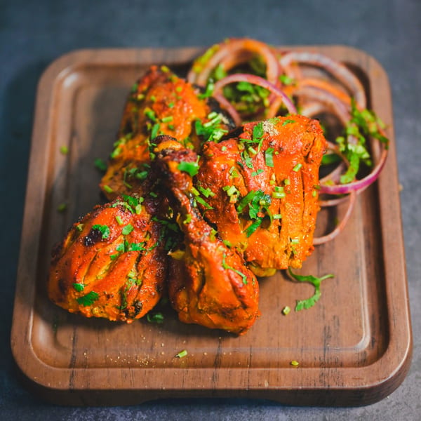 Four pieces of Indian tandoori chicken, garnished with cilantro and red onion, on a wooden board.