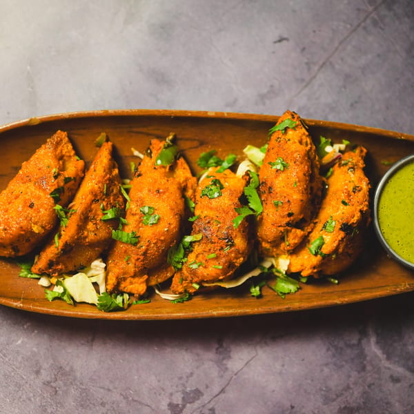 Indian chicken tikka pieces, garnished with cilantro, served with green chutney on a wooden platter.