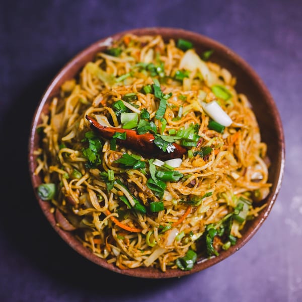 A bowl of Indian noodles with vegetables, green onions, and a red chili pepper.