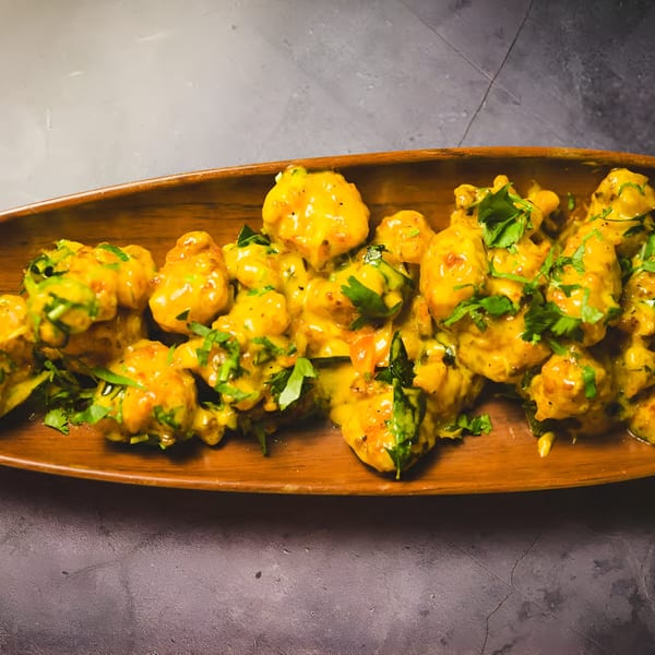 Golden Indian cauliflower florets, coated in a creamy sauce and garnished with fresh green herbs on a wooden platter.